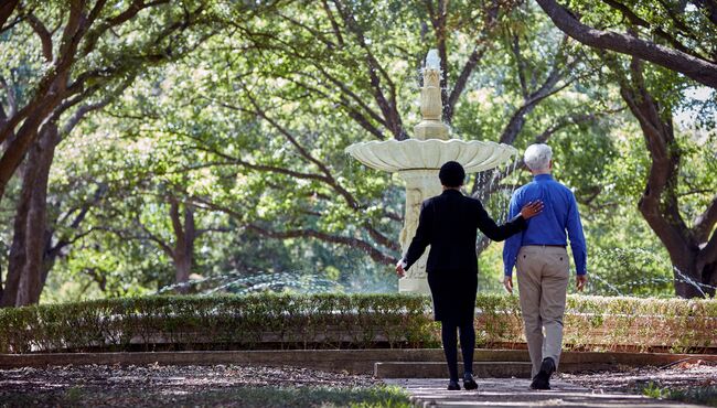 An associate walks a man near a fountain on cemetery grounds.
